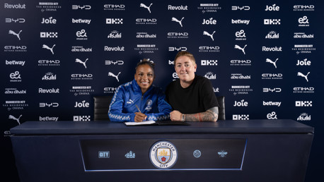 Two people sitting at a desk with Manchester City logos and sponsor background. One is signing a document.
