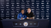 Two people sitting at a desk with Manchester City logos and sponsor background. One is signing a document.