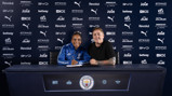 Two people sitting at a desk with Manchester City logos and sponsor background. One is signing a document.