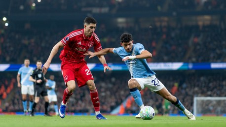 Two soccer players in action during a match. One wears a red kit with number 23, and the other a light blue kit with number 21. They are in a stadium with a blurred referee and players in the background.