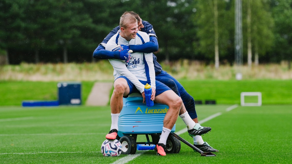 Two football players are having fun on the field during training. One sits on a Lucozade cooler with a water bottle and a soccer ball, while the other playfully hugs him from behind.