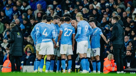 Manchester City players including Reijnders, Matheus Nunes, and Haaland in a huddle on the field during a match with blurred audience in the background.