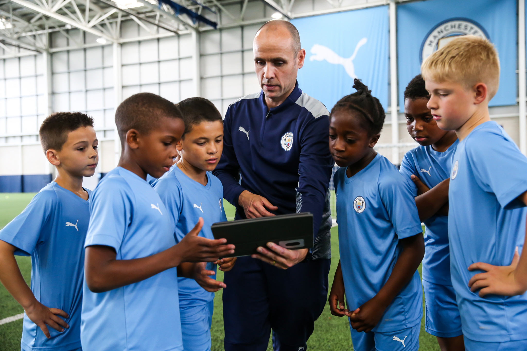 A coach and young players in Manchester City kits indoors at a training session, reviewing tactics on a tablet.