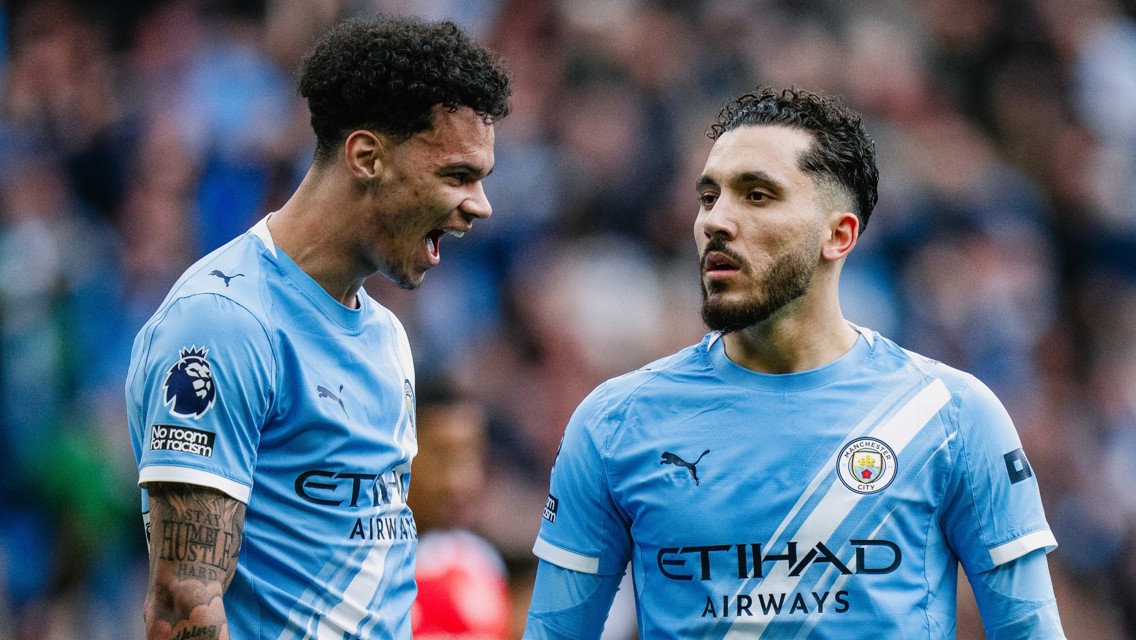 Two soccer players in Manchester City jerseys speaking on the field. Jerseys feature the team logo and sponsor branding.