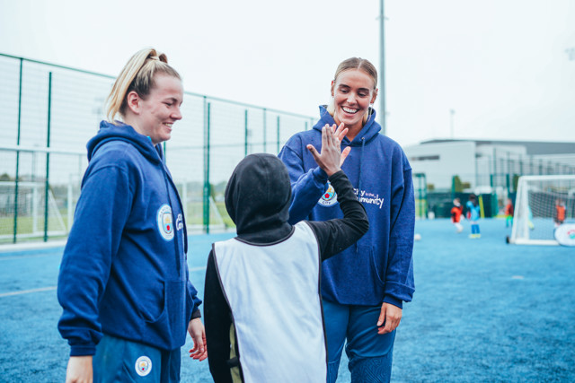 Three people standing on a blue soccer field with two people in blue hoodies, one giving a high-five to another person in a black hoodie and white vest.