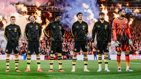 Manchester City players standing in a line wearing black jackets, with a goalkeeper in an orange kit, surrounded by flames in the background.