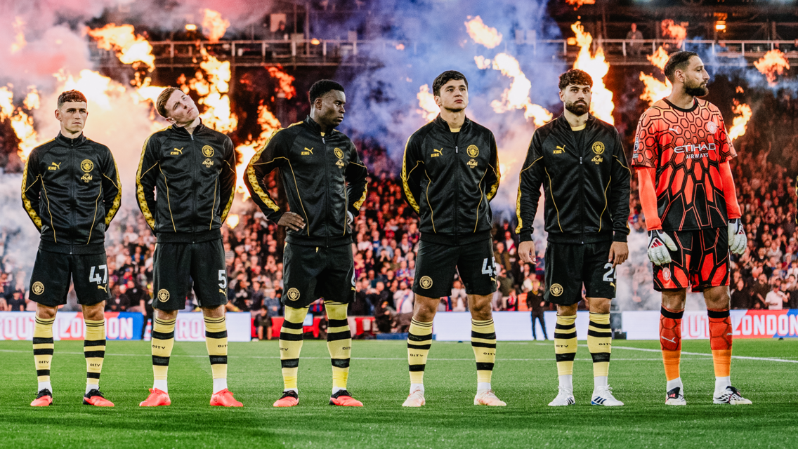 Manchester City players standing in a line wearing black jackets, with a goalkeeper in an orange kit, surrounded by flames in the background.