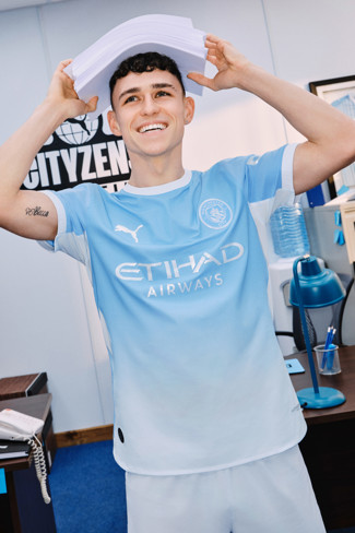A person wearing a Manchester City football jersey holding a stack of papers in an office setting.