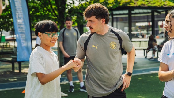 A person wearing a Manchester City shirt interacts with a child at an outdoor event with a Manchester City banner visible.