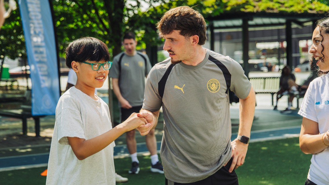 A person wearing a Manchester City shirt interacts with a child at an outdoor event with a Manchester City banner visible.