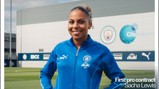 A person in a blue Manchester City tracksuit stands smiling on a field. The background shows the Manchester City logo and the text 'First pro contract Sacha Lewis' displayed on the image.