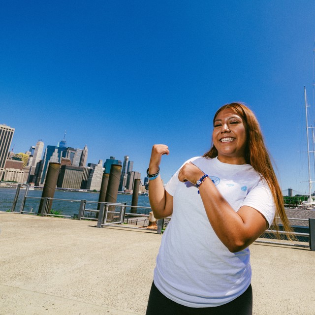 A woman in a white T-shirt is smiling and flexing her arms. Behind her is a view of the Brooklyn Bridge and some skyscrapers in New York City.