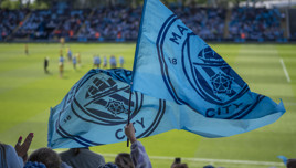 Manchester City fans waving flags at a football match. The flags show the Manchester City logo. The stadium is filled with fans and players are visible on the field.