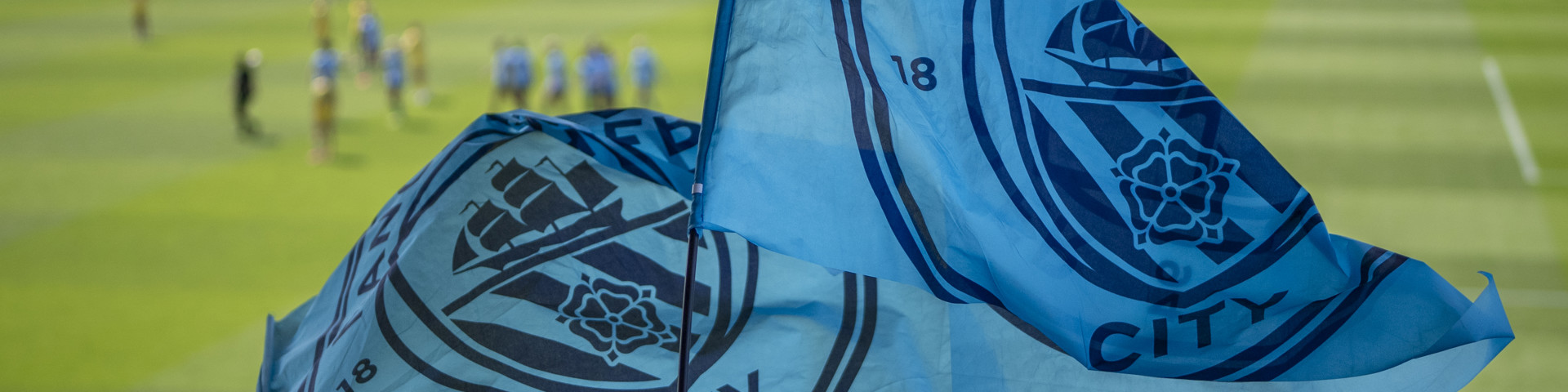 Manchester City fans waving flags at a football match. The flags show the Manchester City logo. The stadium is filled with fans and players are visible on the field.
