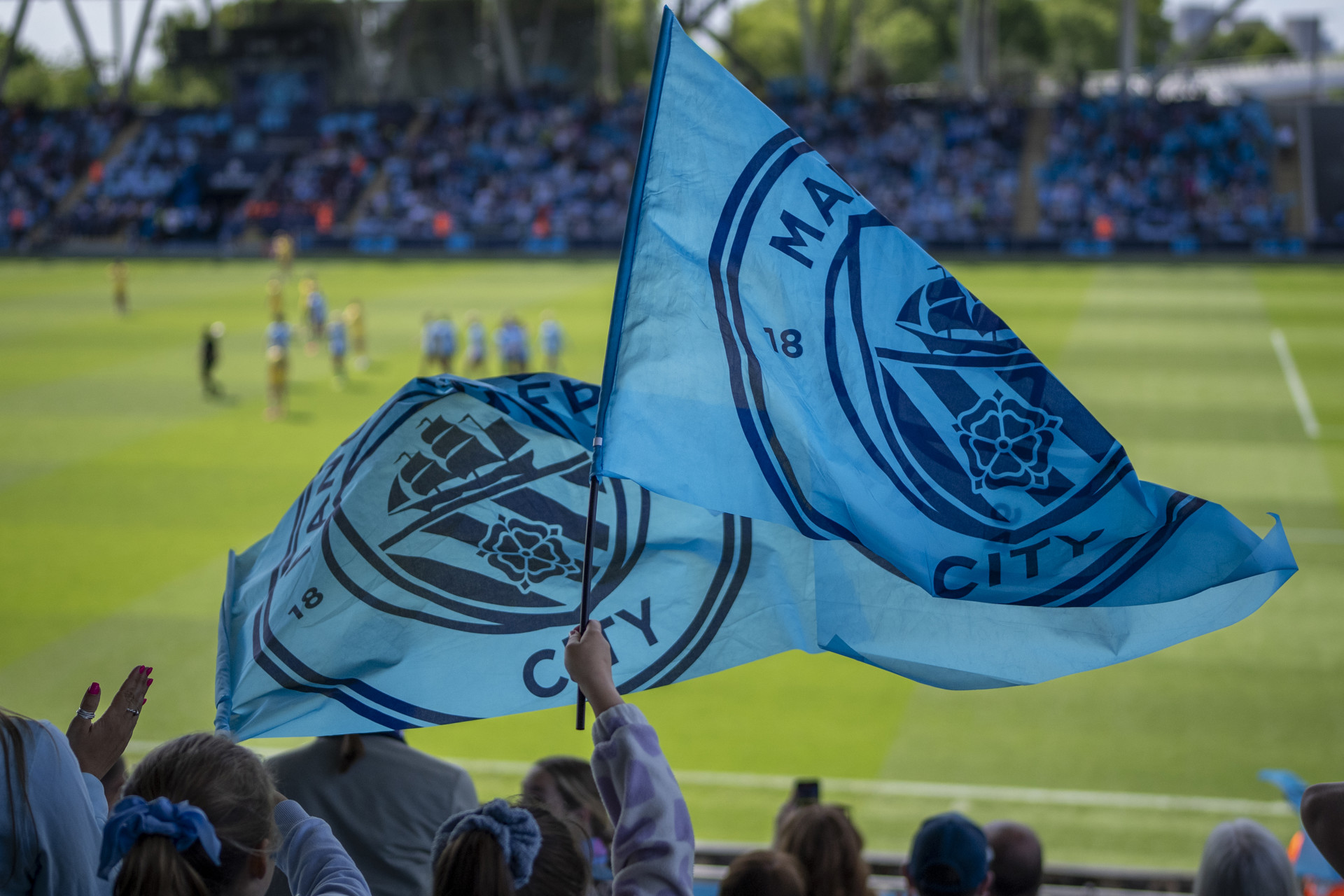 Manchester City fans waving flags at a football match. The flags show the Manchester City logo. The stadium is filled with fans and players are visible on the field.
