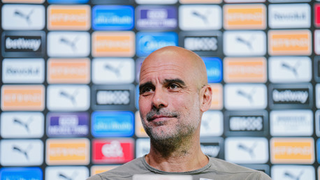 A person with a blurred face at a press conference with a backdrop filled with sponsor logos including Puma and Etihad.