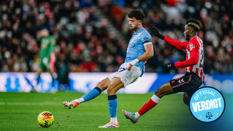 Two soccer players from Manchester City and Southampton are in action during a match. The Manchester City player is wearing a light blue kit, while the Southampton player is in a red and white striped kit. The image includes an 'Official Verdict' emblem.