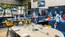 Dining area with Manchester City Women's team posters, plants, tables set with cutlery and bottles, and screens showing a football match.