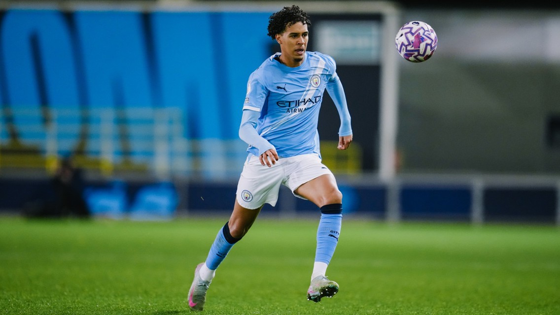 A Manchester City player in action wearing the team's light blue jersey and white shorts, running towards a soccer ball on a field.