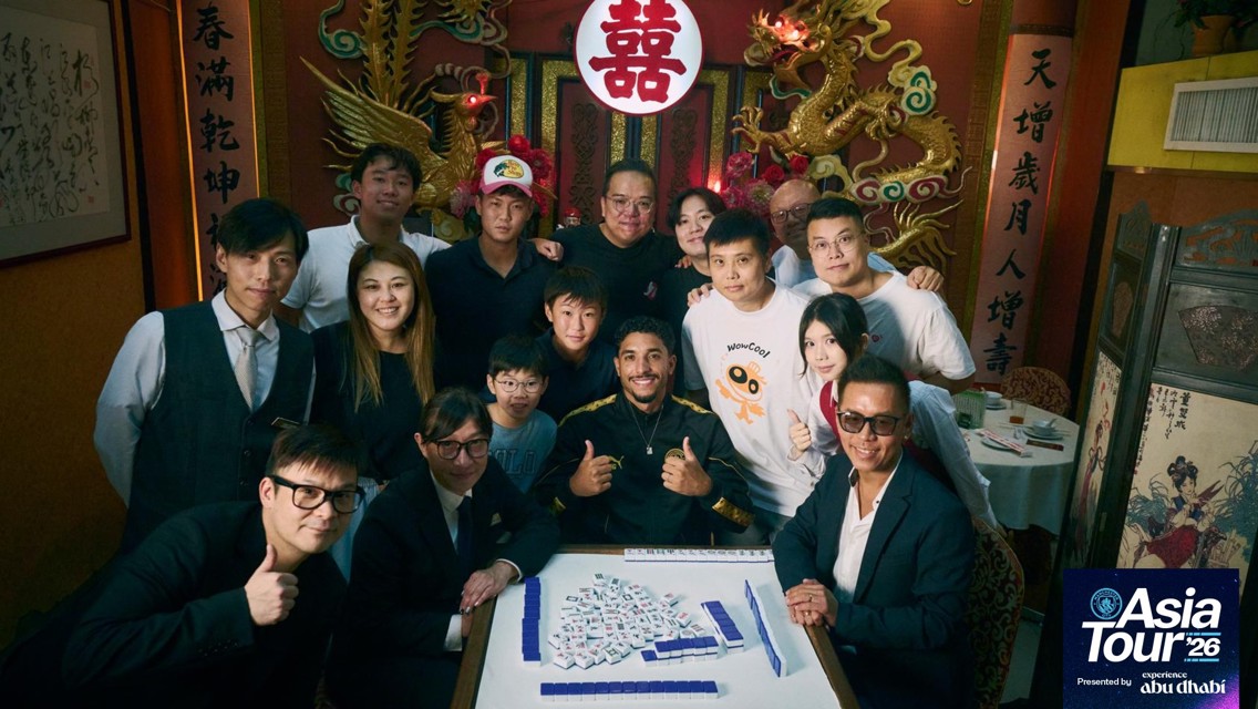 A group of people posing around a table with mahjong tiles in a decoratively adorned room, featuring Chinese symbols and decorations. A logo for 'Asia Tour '26' is visible at the bottom right.