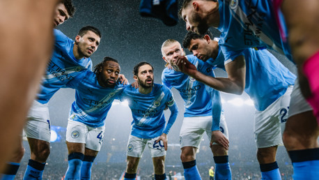 A group of Manchester City players in light blue jerseys, huddling together on the field during a match, with blurred faces. The Etihad Stadium is visible in the background under lights.