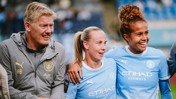 Three individuals, including two wearing Manchester City women's football kits and a coach, smiling and posing together on the field.