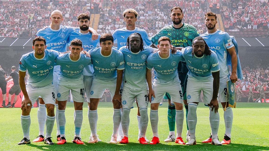 A group of Manchester City football players posing on a field for a team photo. They are wearing light blue jerseys sponsored by Etihad Airways with the logo visible. A crowd is in the background.