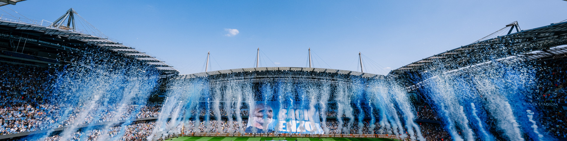 A football match celebration at a stadium with confetti and flags. The field displays a large Manchester City crest.