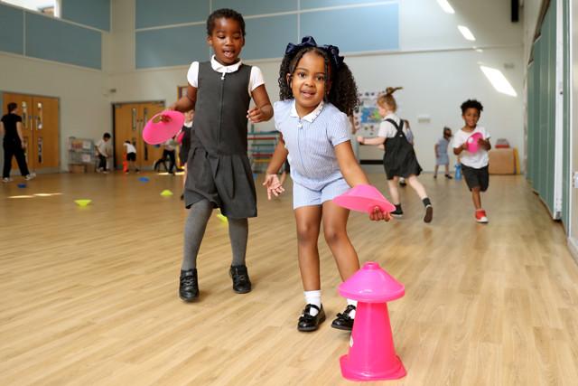 Young children in a gym setting are participating in a game involving colorful plastic cones.