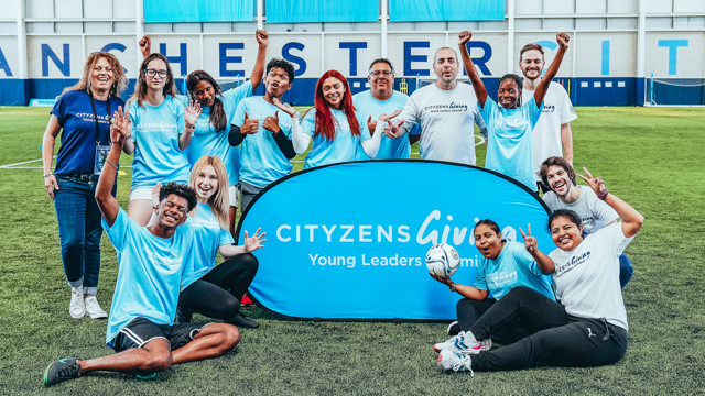 A group photo at a Cityzens Giving Young Leaders Summit event in a Manchester City football facility. The attendees are wearing light blue shirts and are posing in front of a Cityzens banner.