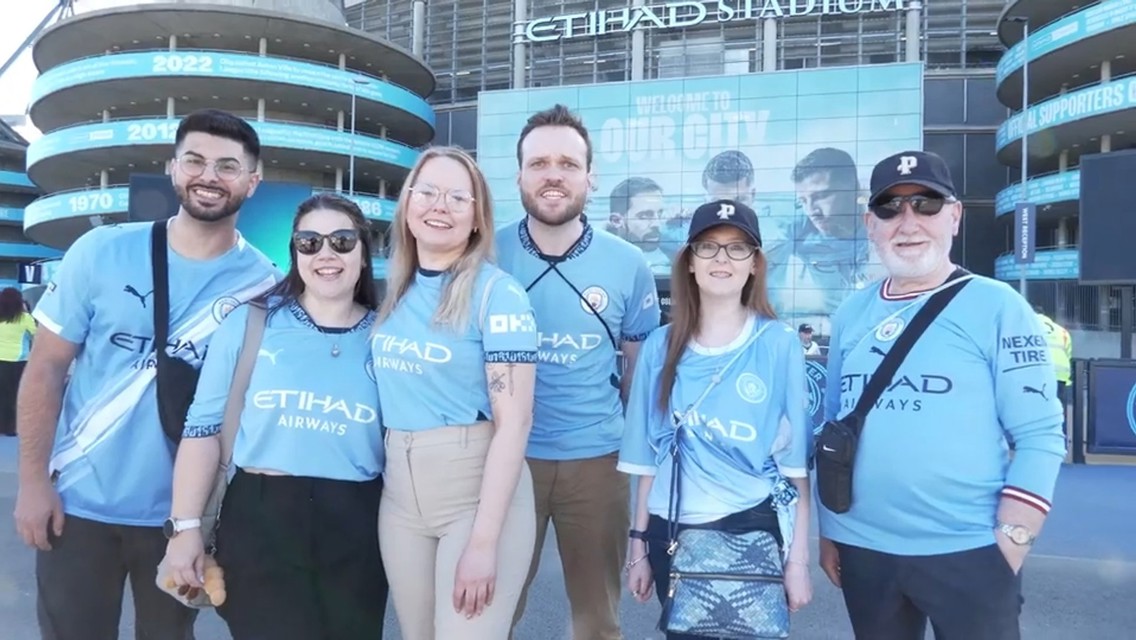 A group of people wearing Manchester City jerseys posing in front of the Etihad Stadium.