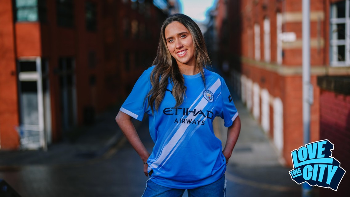 A person wearing the Manchester City football kit with Etihad Airways sponsorship stands in an urban area. The Manchester City crest is visible on the jersey. A 'Love This City' logo appears in the bottom right corner.