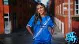 A person wearing the Manchester City football kit with Etihad Airways sponsorship stands in an urban area. The Manchester City crest is visible on the jersey. A 'Love This City' logo appears in the bottom right corner.