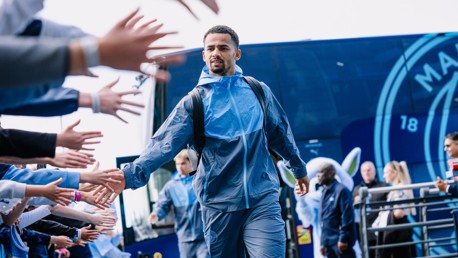 A football player wearing a blue tracksuit greets fans by high-fiving as he walks past them. Behind him is a blue Manchester City bus.