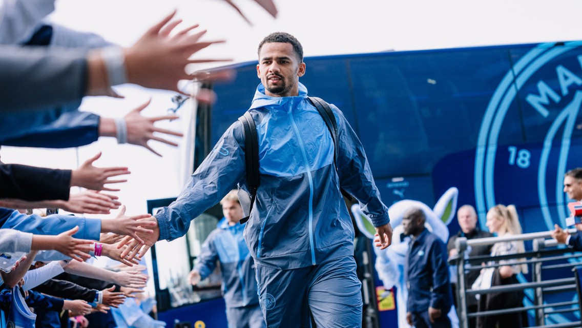 A football player wearing a blue tracksuit greets fans by high-fiving as he walks past them. Behind him is a blue Manchester City bus.