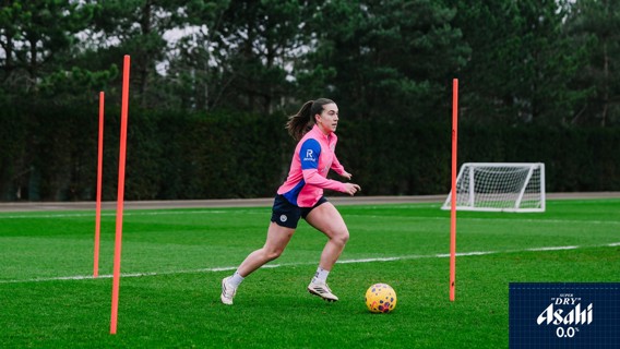 A football player in a pink and blue kit practices dribbling through orange poles on a grassy field with a goalpost in the background.