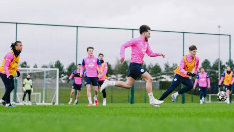 Soccer players in pink jerseys are actively training on a field, with one player chasing a ball. A goalkeeper and the goalpost are visible in the background.