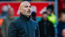 A person in a black jacket stands at a football stadium with a blurred crowd in the background.