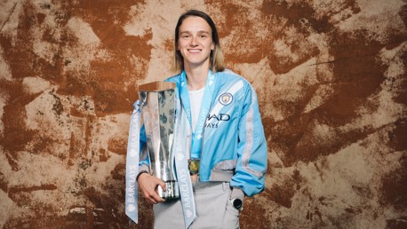 A person wearing a Manchester City jacket holding a Barclays trophy, with a medal around their neck.