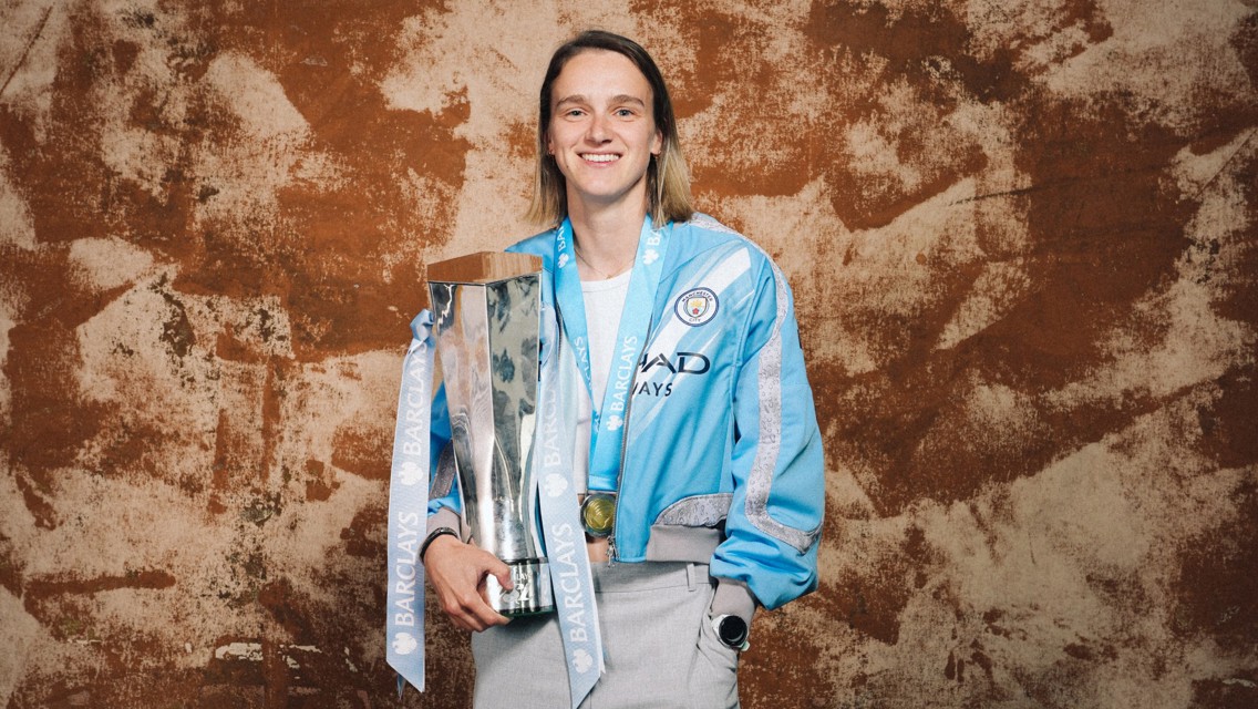 A person wearing a Manchester City jacket holding a Barclays trophy, with a medal around their neck.