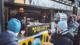 A crowd gathers in front of the 'Northern Soul Grilled Cheese' food truck. People are dressed in winter attire, holding plates of food, as a vendor hands over orders at the 'Collect' counter.