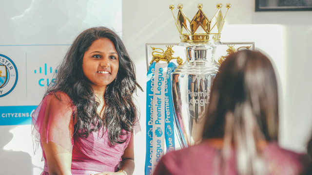 A person in a maroon dress is posing near the Premier League trophy, which is adorned with blue ribbons and the Manchester City logo visible in the background.
