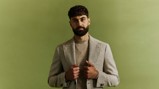A man with a beard wearing a houndstooth sport coat over a beige shirt, against a light green background.