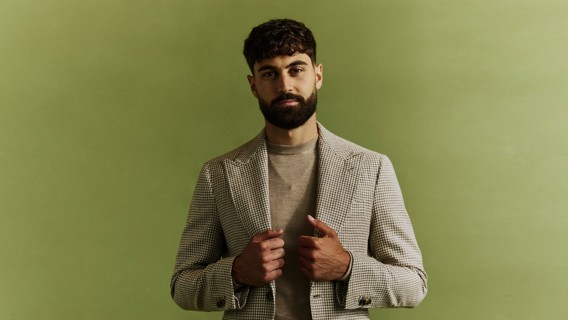 A man with a beard wearing a houndstooth sport coat over a beige shirt, against a light green background.