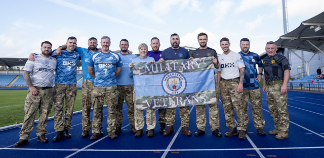 A group of military veterans dressed in camouflage pants and various Manchester City jerseys posing on a track while holding a flag that reads 'MILITARY VETERANS Manchester City'.