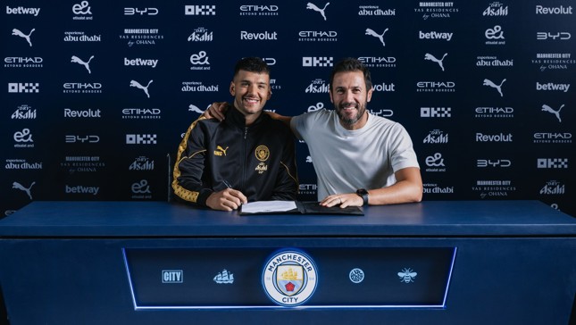 Two individuals sitting at a table with Manchester City branding, signing a contract. The background displays various sponsor logos such as Puma, Betway, Etihad, and Revolut.