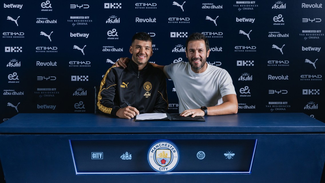 Two individuals sitting at a table with Manchester City branding, signing a contract. The background displays various sponsor logos such as Puma, Betway, Etihad, and Revolut.