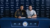 Two individuals sitting at a table with Manchester City branding, signing a contract. The background displays various sponsor logos such as Puma, Betway, Etihad, and Revolut.