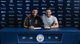 Two individuals sitting at a table with Manchester City branding, signing a contract. The background displays various sponsor logos such as Puma, Betway, Etihad, and Revolut.