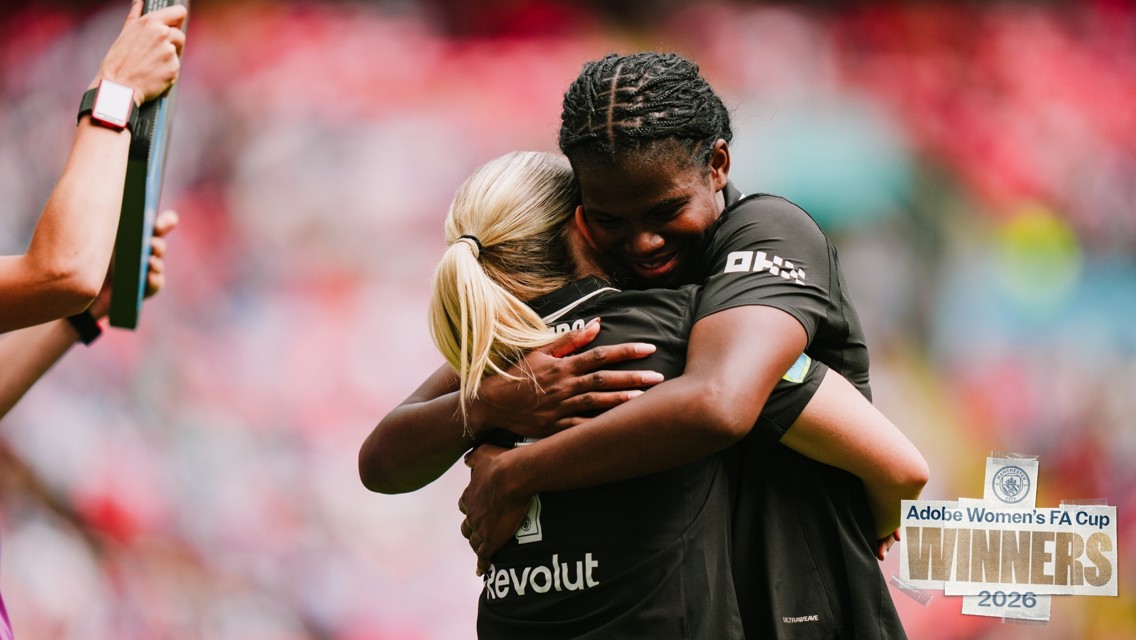 Two athletes in black uniforms embrace on a sports field, celebrating with a banner reading 'Adobe Women's FA Cup Winners 2026' in the foreground.